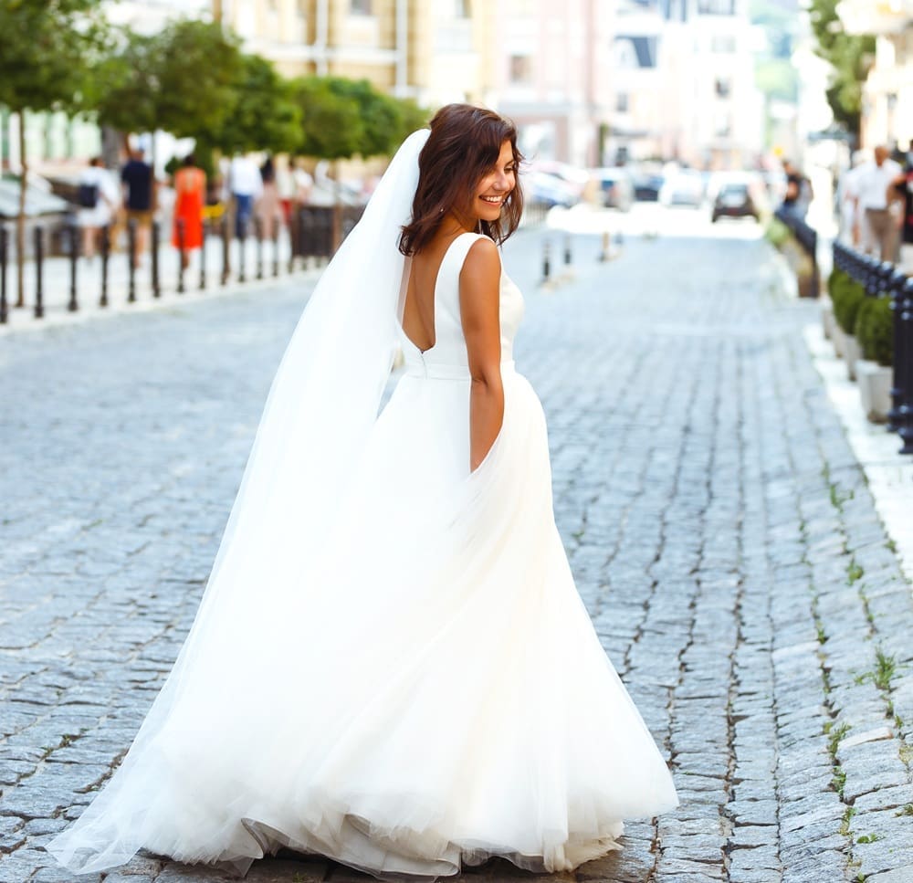 A bride in a white wedding dress and veil stands on a cobblestone street, looking back and smiling, with buildings and people in the background.