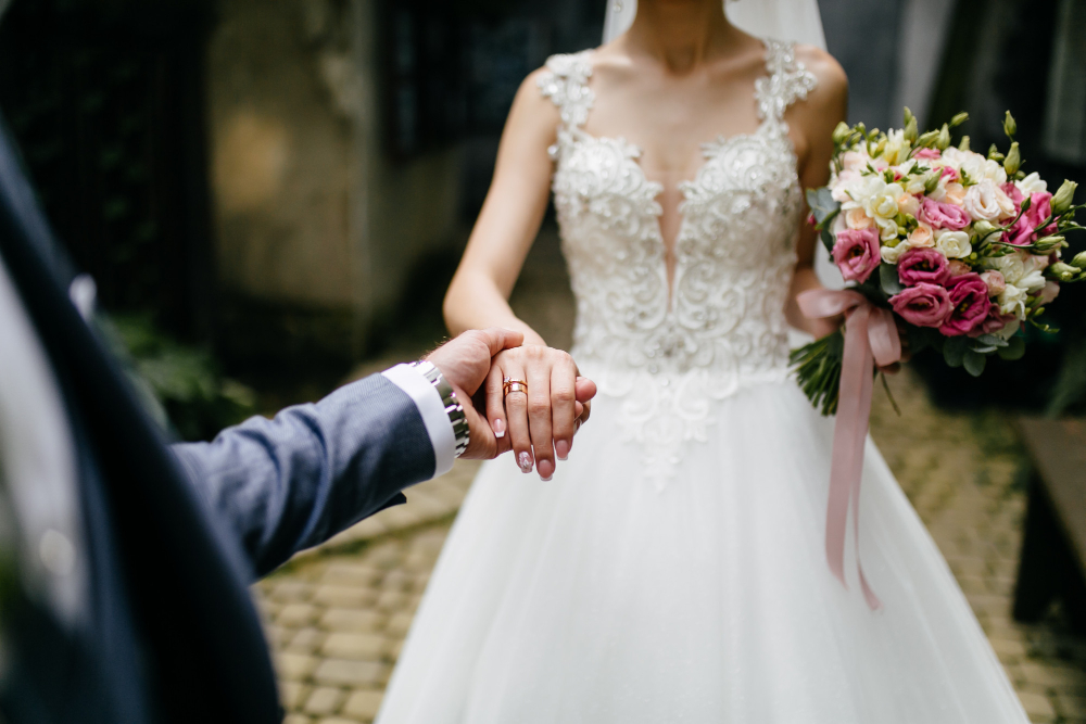 A bride in a white wedding dress holding a bouquet of pink flowers clasps hands with someone in a suit.