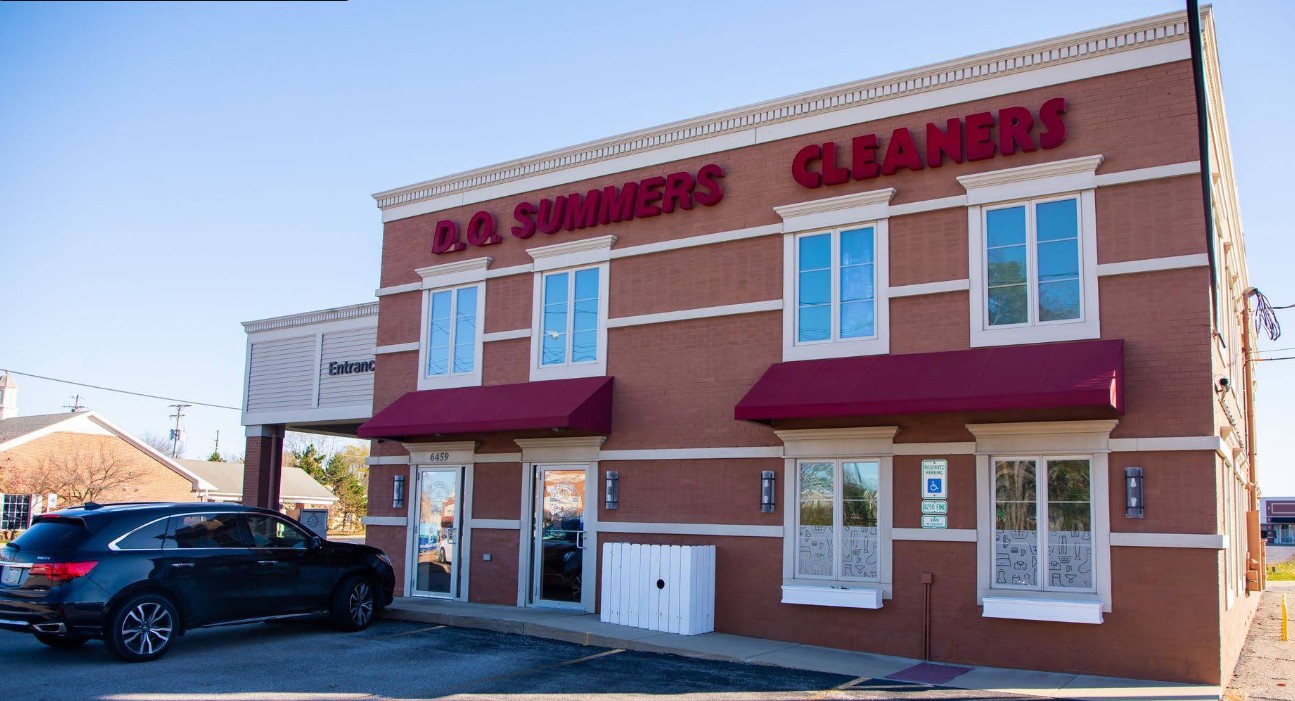 A brick building with red awnings and large lettering reading “D.C. Summers Cleaners.” A black SUV is parked in front near the entrance.