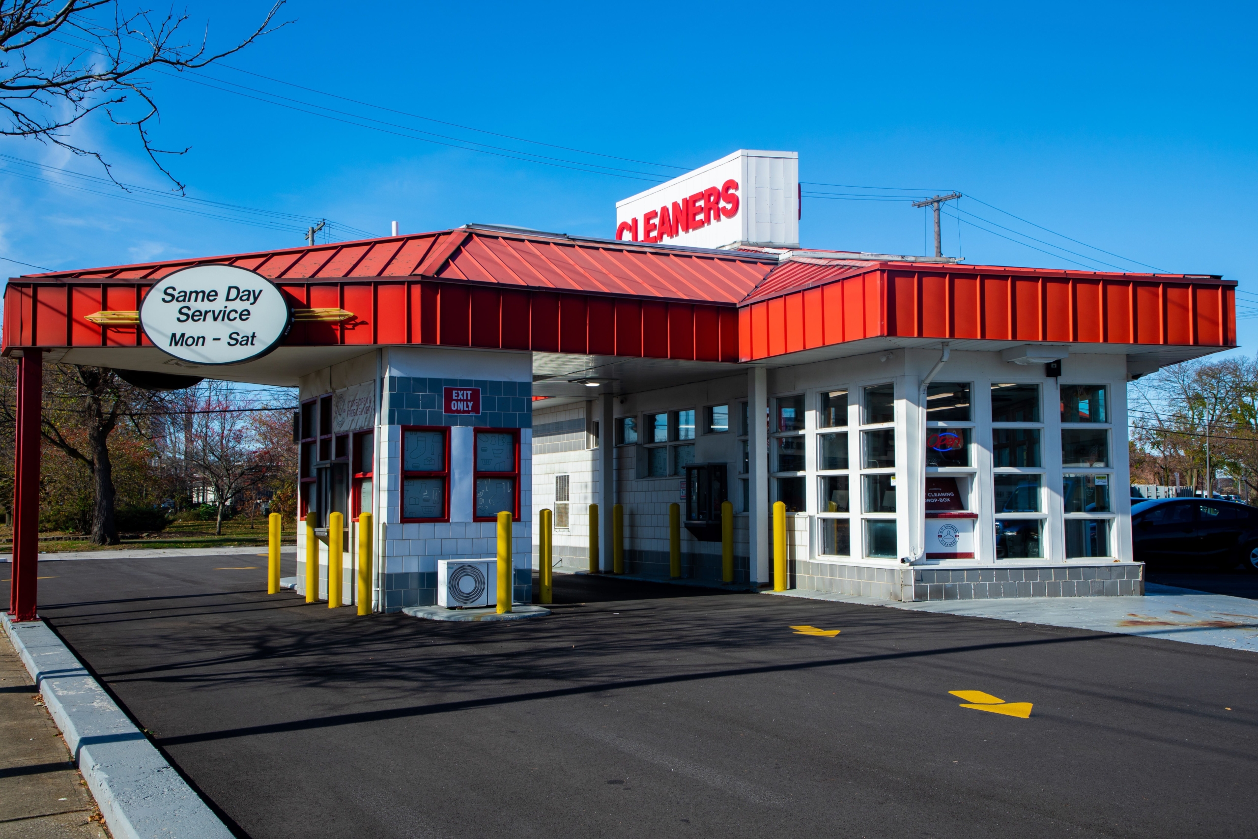 A drive-thru dry cleaners building with a red roof, white walls, and a sign offering same-day service from Monday to Saturday.