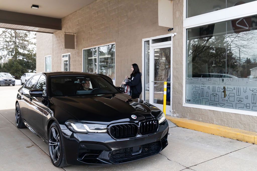 A black BMW sedan is parked in front of a commercial building while a person stands near the entrance door holding items.