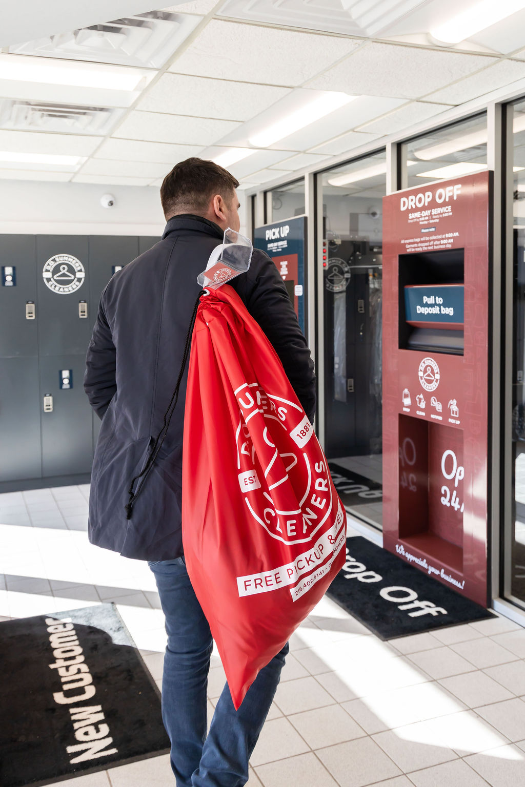A man carrying a red dry cleaning bag walks toward self-service lockers and a drop-off screen in a modern dry cleaner's facility.