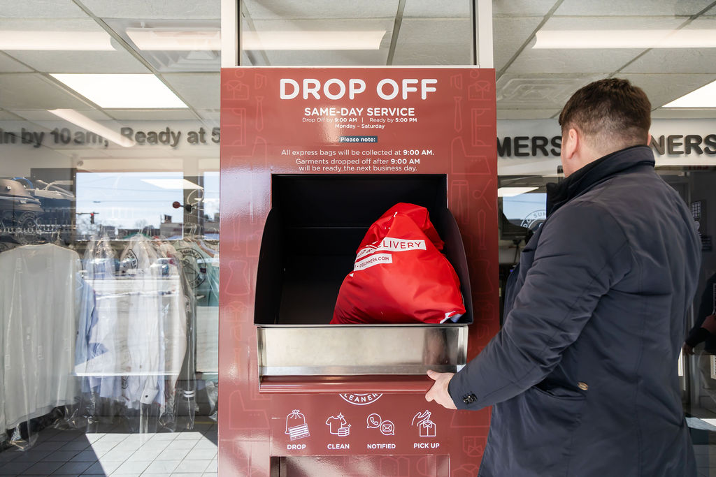 A person places a red laundry bag into a drop-off slot at a dry cleaning service counter with same-day service information displayed above.