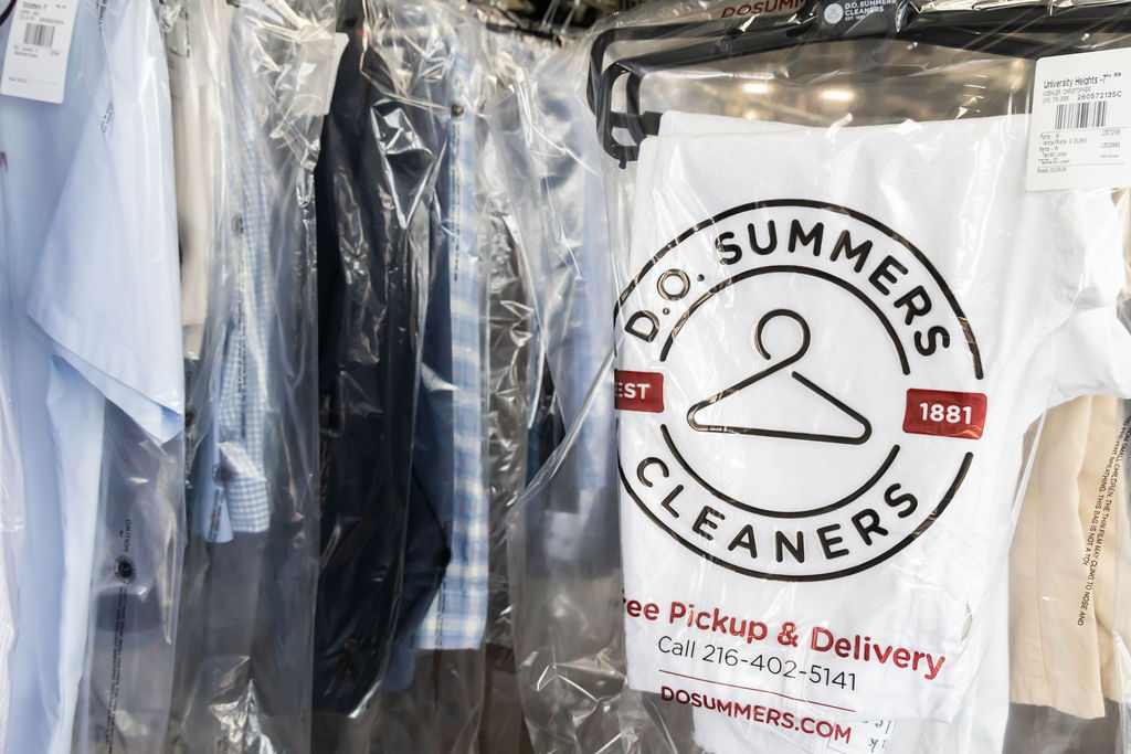 Clothes covered in plastic are hanging on a rack at a dry cleaner, with a D.O. Summers Cleaners bag and logo visible in the foreground.