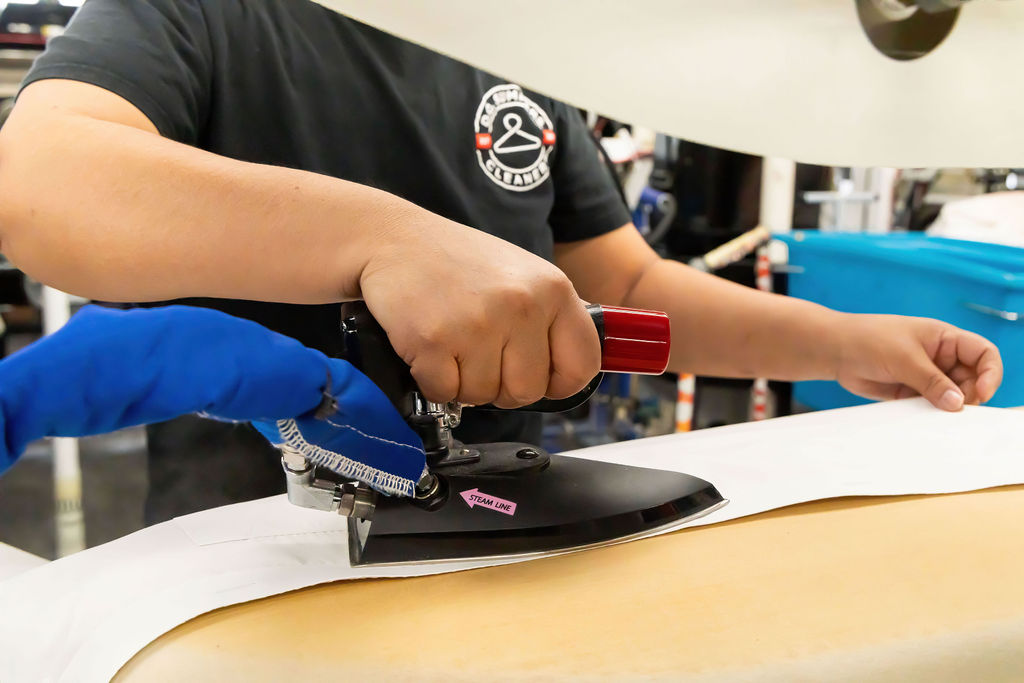 A person uses a steam iron to press a white garment on an ironing board, assisted by another person wearing a blue glove.