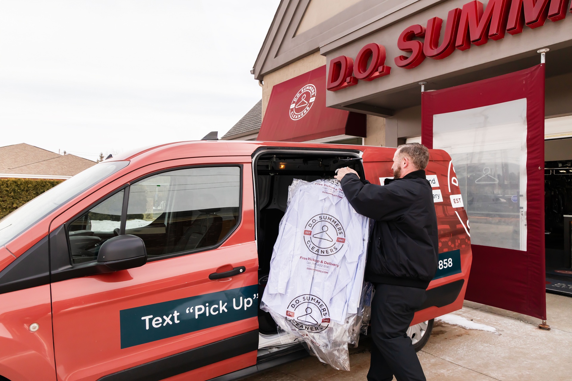 A man loads dry cleaned clothes covered in plastic into a red D.O. SUMMIT van parked outside the store.